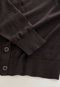 Tumši brūns garais piedurkņu kardigāns ar ribotām aprocēm un apakšmalu, kam priekšējā atvērumā ir divi lieli pogas.