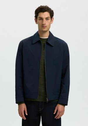 Homme aux cheveux courts et bouclés portant une veste zippée bleu marine sur un pull vert et un jean foncé, debout devant un fond clair uni.