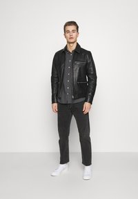 Chaqueta de cuero negra con una textura suave, cremallera frontal y dos bolsillos con cremallera, llevada sobre una camiseta gris y combinada con jeans oscuros y zapatillas blancas.