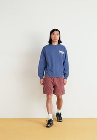 Sweatshirt azul em algodão com gola redonda e logótipo, combinado com calções de corduroy rosa. Ténis pretos e meias brancas completam o visual.