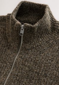 Bruine gebreide trui met een hoge halslijn en een zilveren rits. Gestructureerde stof met een grove, warme uitstraling.