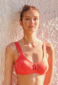 Jeune femme aux cheveux attachés portant un haut de bikini corail, debout contre un mur clair texturé dans une lumière naturelle douce.