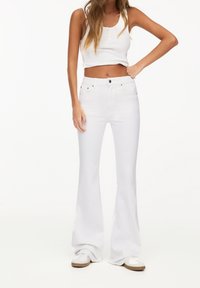 Jeans blancos acampanados de mezclilla con una cintura ajustada y sutiles bolsillos frontales. Combinados con una camiseta sin mangas blanca y zapatillas blancas.