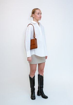 Femme portant une chemise blanche oversize, une mini-jupe beige, des bottes noires montantes et un sac à épaule marron, debout devant un fond blanc.