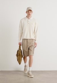 Camisa polo de mangas compridas beje, calções claros de cor areia com bolsos laterais, sapatilhas creme, e um boné a condizer; a segurar uma mala tote de cor areia.