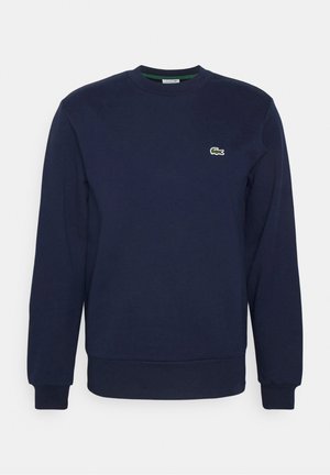 Persoon draagt een marineblauwe trui, groene joggingbroek, zwarte sneakers met blauwe veters en houdt een marineblauwe rugzak vast tegen een effen achtergrond.
