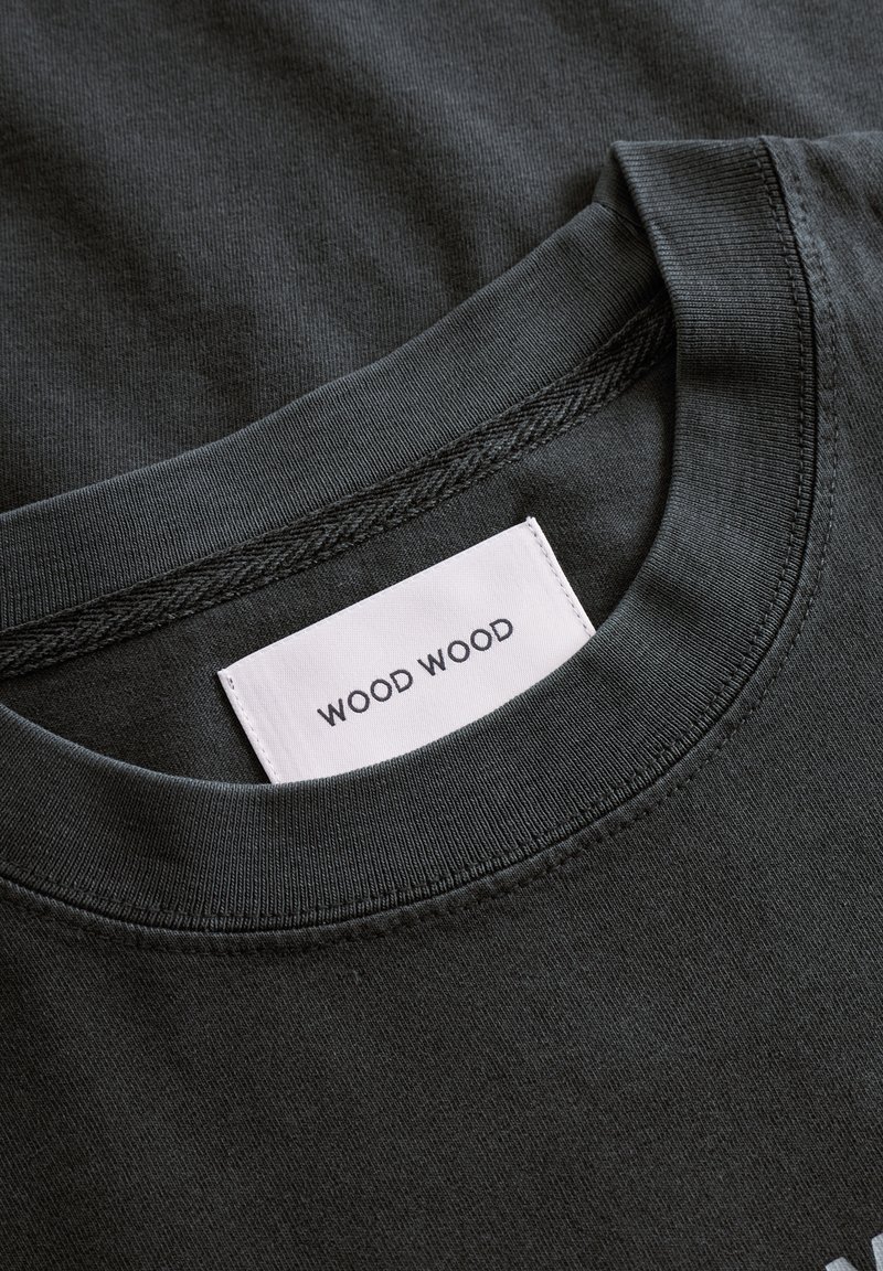T-shirt en coton gris foncé avec un col rond et une étiquette blanche avec le texte "WOOD WOOD" cousu à l'intérieur du col.