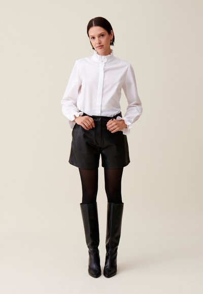 Chemise blanche à col froncé et à poignets volantés, assortie à un short en cuir noir, des collants noirs et des bottes noires à talons hauts jusqu'aux genoux.
