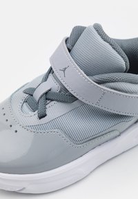 Šedá sportovní obuv s lesklým syntetickým svrškem, síťovanými akcenty a polstrovaným límcem. Obsahuje texturovaný pásek a šedé tkaničky.