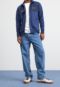 Marinblå zip-up jacka med svarta detaljer, vit t-shirt, blå denimjeans och vita sneakers med marinblå detaljer. Har Nike-logotyper.