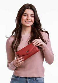 Červená kožená peněženka s texturovaným vzorem a embosovaným logem, s klopovým uzávěrem. Model ji drží v růžovém svetru na bílém pozadí.
