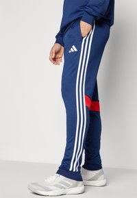 Navy joggers with three white stripes along the sides, red accent near the hem, and an elastic waistband. Paired with white sneakers.