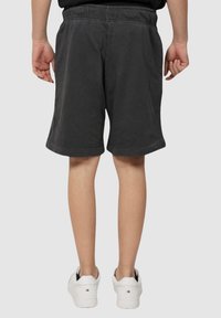 Person in schwarzen knielangen Shorts und weißen Sneakers, stehend mit entspannten Händen an den Seiten, von hinten gesehen.