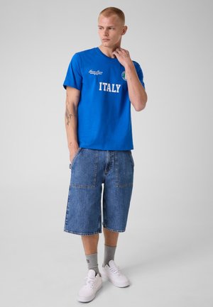 Joven con el pelo corto, vestido con una camiseta azul con la palabra "ITALY", pantalones vaqueros cortos hasta la rodilla, calcetines grises y zapatillas blancas, de pie frente a un fondo liso.