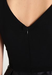 Svart klänning med en djup V-ringning, slät tyg och osynlig dragkedja bak. Texturerad silhuett med en figursydd överdel.