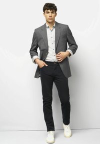 Ein junger Mann steht an einer weißen Wand, trägt ein graues Blazer über einem hellgrauen Hemd, schwarze Hose und weiße Sneakers, mit einer Hand in der Tasche.