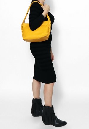 Vrouw in een zwarte jurk en zwarte enkellaarsjes met franjes, draagt een gele schoudertas met gevlochten handvat en staat zijwaarts.