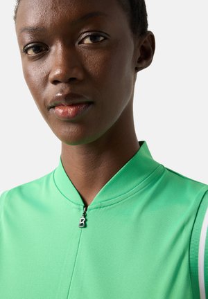 Κοντινό πλάνο ενός ατόμου με σκουρόχρωμο δέρμα που φοράει αμάνικο ανοιχτό πράσινο τοπ με μικρό φερμουάρ και τραβηχτήρι φερμουάρ με το γράμμα "B".