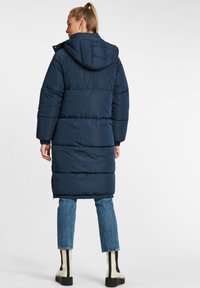 Navy puffer coat with a hood, quilted design, long length, and ribbed cuffs, paired with light denim and black and white heeled boots.