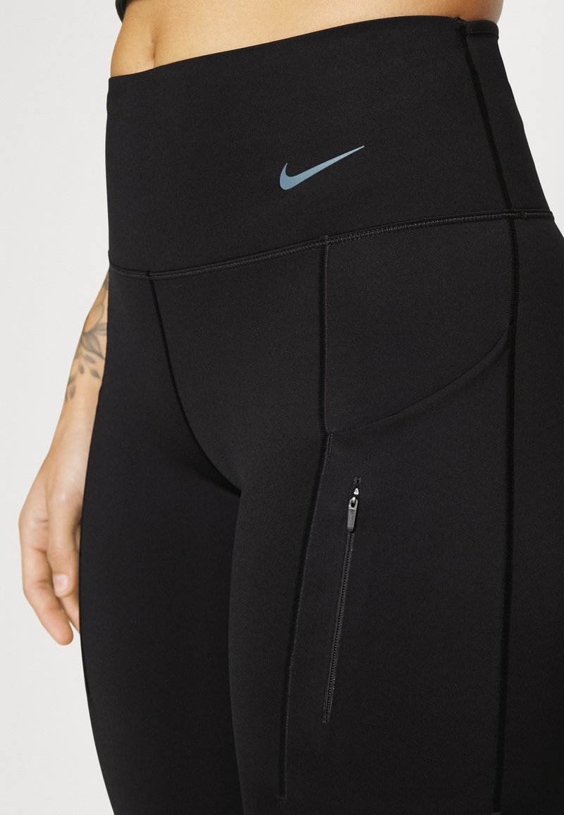 Gros plan sur une personne portant des leggings taille haute noirs de Nike avec une poche zippée sur le côté et un logo swoosh Nike bleu clair.