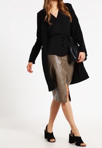 Haut noir portefeuille avec une taille ceinturée, jupe crayon métallisée à motif crocodile avec une fente devant, et sandales à talons carrés noires.