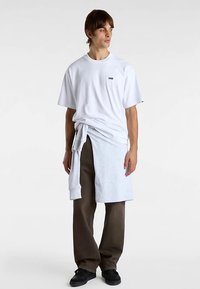 Vit t-shirt med korta ärmar och en liten svart logotyp, lager-på-lager med en ljusgrå sweatshirt knuten i midjan, kombinerad med lösa bruna byxor och svarta skor.