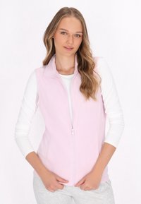 Gilet in pile rosa chiaro con colletto alto, chiusura frontale con zip e due tasche laterali. Abbinato a una maglietta bianca a maniche lunghe e pantaloni grigi.