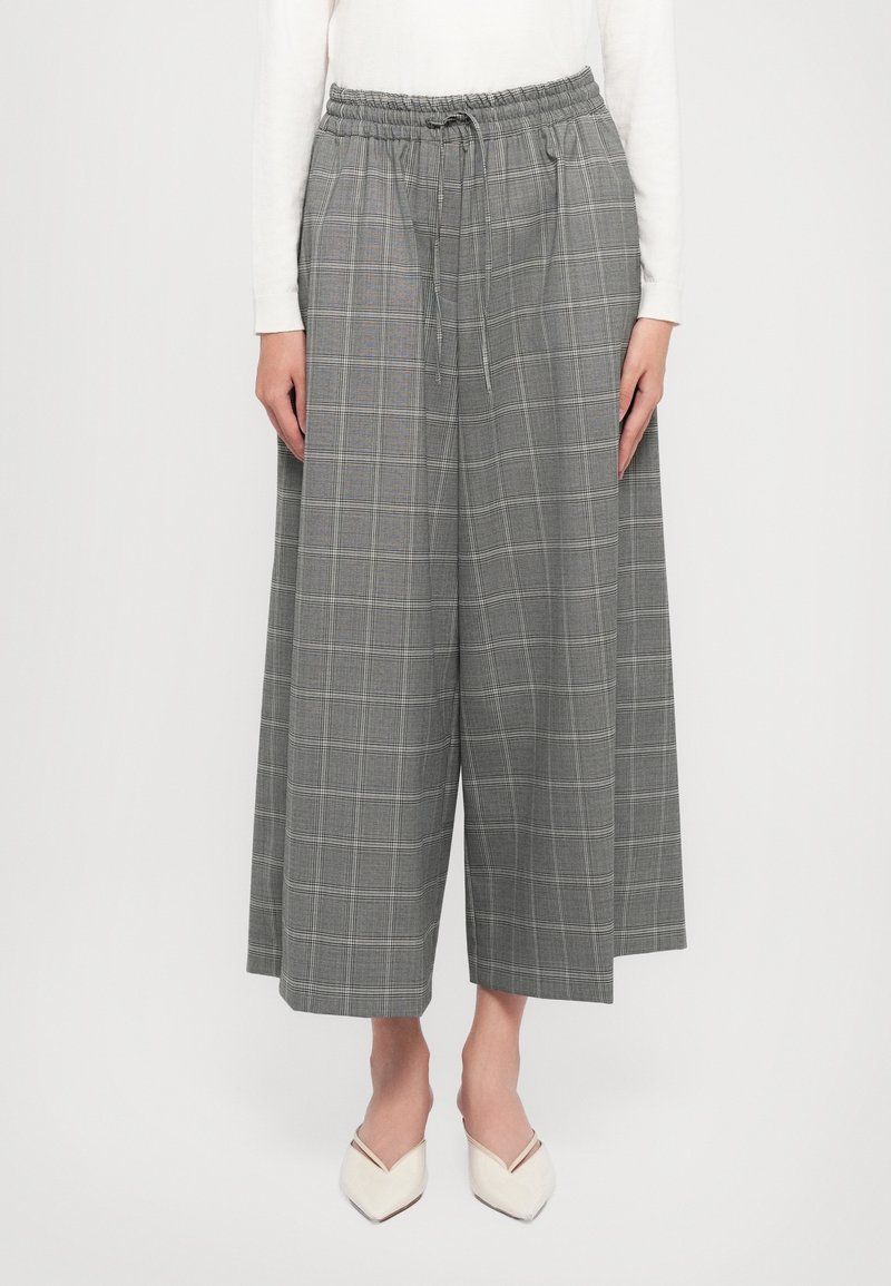 Pantalon gris à jambe large et raccourci avec un motif à carreaux subtil, taille élastique avec cordon, porté avec des chaussures blanches pointues et un haut blanc.