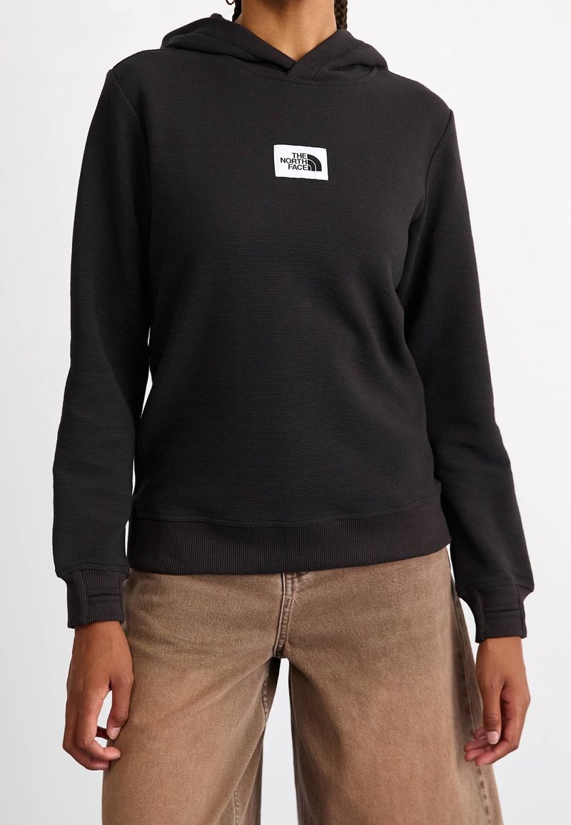 Felpa nera con cappuccio realizzata in tessuto strutturato, con un patch logo bianco sul petto, polsini e orlo a costine, abbinata a pantaloni larghi color marrone chiaro.