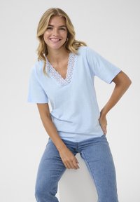Camiseta de manga corta azul claro con escote en V que presenta un delicado borde de encaje blanco. Combinada con jeans de mezclilla azul, la tela se ve suave y casual.