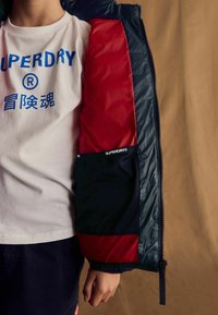 Chaqueta puffer azul marina con acentos rojos y tela texturada negra, con cremallera al frente, llevada sobre una camiseta gráfica blanca.
