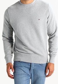 Sudadera gris de cuello redondo hecha de un tejido suave, con puños y dobladillo acanalados, y un pequeño logo bordado en el pecho. Combinada con unos jeans azules.