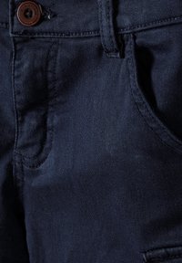 Tmavě modrá denimová látka s viditelným šitím, kapsami a hnědým knoflíkem. Hladká textura s mírným leskem, detailní pohled na materiál.