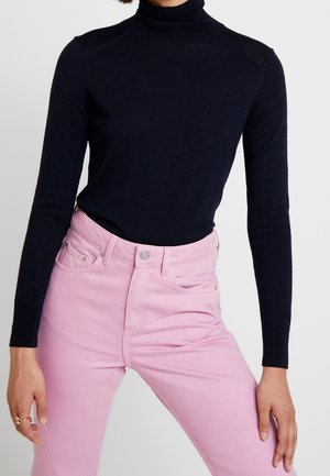 Femme portant un pull col roulé noir ajusté rentré dans un jean taille haute rose clair, se tenant debout avec les bras détendus.