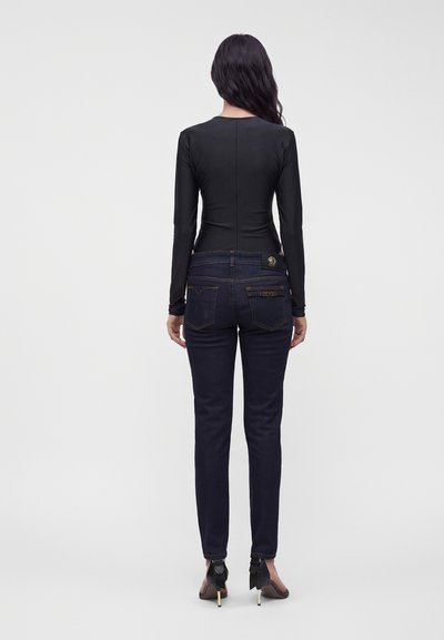 Femme aux longs cheveux foncés portant un haut noir cintré à manches longues, un jean bleu foncé et des chaussures à talons noirs, debout de dos.