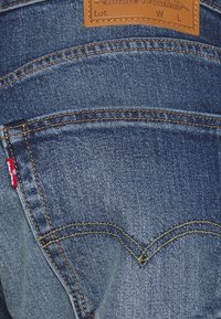 Modré džíny z denimu s koženou zadní nášivkou, charakteristickým obloukovým stehem na kapsách a malou červenou logovou visačkou na boku.