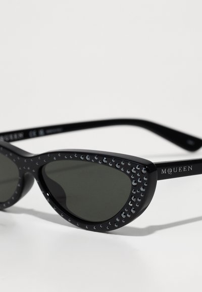 Ochelari de soare negri cu formă de ochi de pisică, cu lentile întunecate, împodobiți cu mici cristale negre în jurul ramei și cu "McQueen" pe brațul templei.