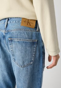 Ljusblå denimjeans med en slitagen textur, med en brun läderlapp som är märkt "Calvin Klein Jeans" på midjebandet.