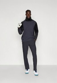 Golfista maschio con un abbigliamento sportivo scuro e guanti da golf bianchi, che tiene una mazza da golf sulle spalle, in piedi contro uno sfondo neutro.