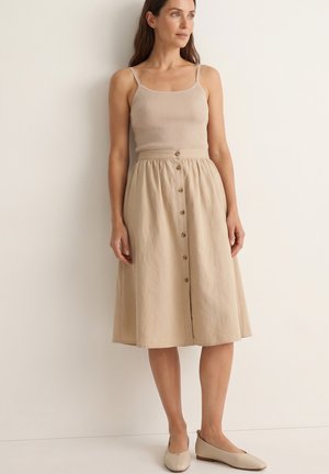 Kvinde iført en beige ribbet tanktop, beige midi-nederdel med høj talje og knapper samt beige flade sko, der står foran en ensartet lys væg.