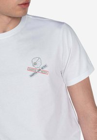 Camiseta de algodón blanca con un pequeño gráfico de una calavera con huesos cruzados, superpuesto con texto en rojo y gris que dice "HARPER Y NUNCA".