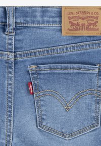 Los vaqueros de mezclilla azul claro presentan un bolsillo trasero distintivo con costuras doradas y una etiqueta roja. La tela tiene una textura suave con patrones típicos.