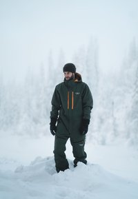 Zielony zimowy kombinezon z pomarańczowymi akcentami, kapturem i zamykanymi na zamek kieszeniami. Ubrany z czarnymi rękawiczkami i dzianą czapką, stojący na śniegu.