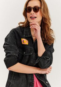 Czarna kurtka dżinsowa z podwiniętymi rękawami, ozdobiona kolorową naszywką. Pod nią częściowo widoczny różowy top o fakturze. Na nosie brązowe, okrągłe okulary przeciwsłoneczne.