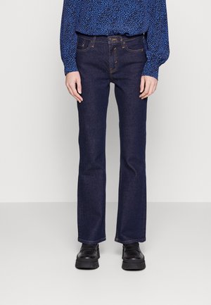 Persoon draagt donkere blauwe recht gesneden jeans, blauw patroon langarmshirt en zwarte grove schoenen, staand tegen een effen achtergrond.