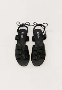 Mustad mustad sandaalid, millel on punutud nahkdisain, avatud varvas ja pahkluu sidemed. Omavad pehmet tekstuuri ja minimalistlikke riistvaraelemente.