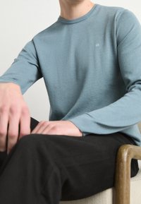 Jasnoniebieski sweter z długim rękawem i okrągłym dekoltem, o prążkowanej fakturze, z wyszytym logo "ck". W połączeniu z czarnymi spodniami, ręce opierają się na kolanach.