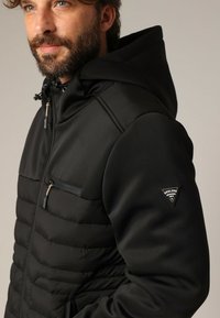 Homme avec une barbe portant une veste matelassée noire avec capuche et poche zippée sur la poitrine, logo de la marque Deeluxe sur la manche gauche.