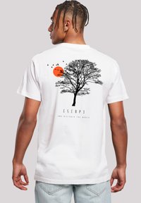 Mann mit geflochtenem Haar trägt ein weißes T-Shirt mit schwarzem Baum, fliegenden Vögeln, orangefarbenem Sonnenmotiv und dem Text "ESCAPE" auf dem Rücken.