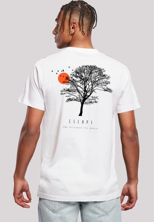 Mann mit geflochtenem Haar trägt ein weißes T-Shirt mit schwarzem Baum, fliegenden Vögeln, orangefarbenem Sonnenmotiv und dem Text "ESCAPE" auf dem Rücken.